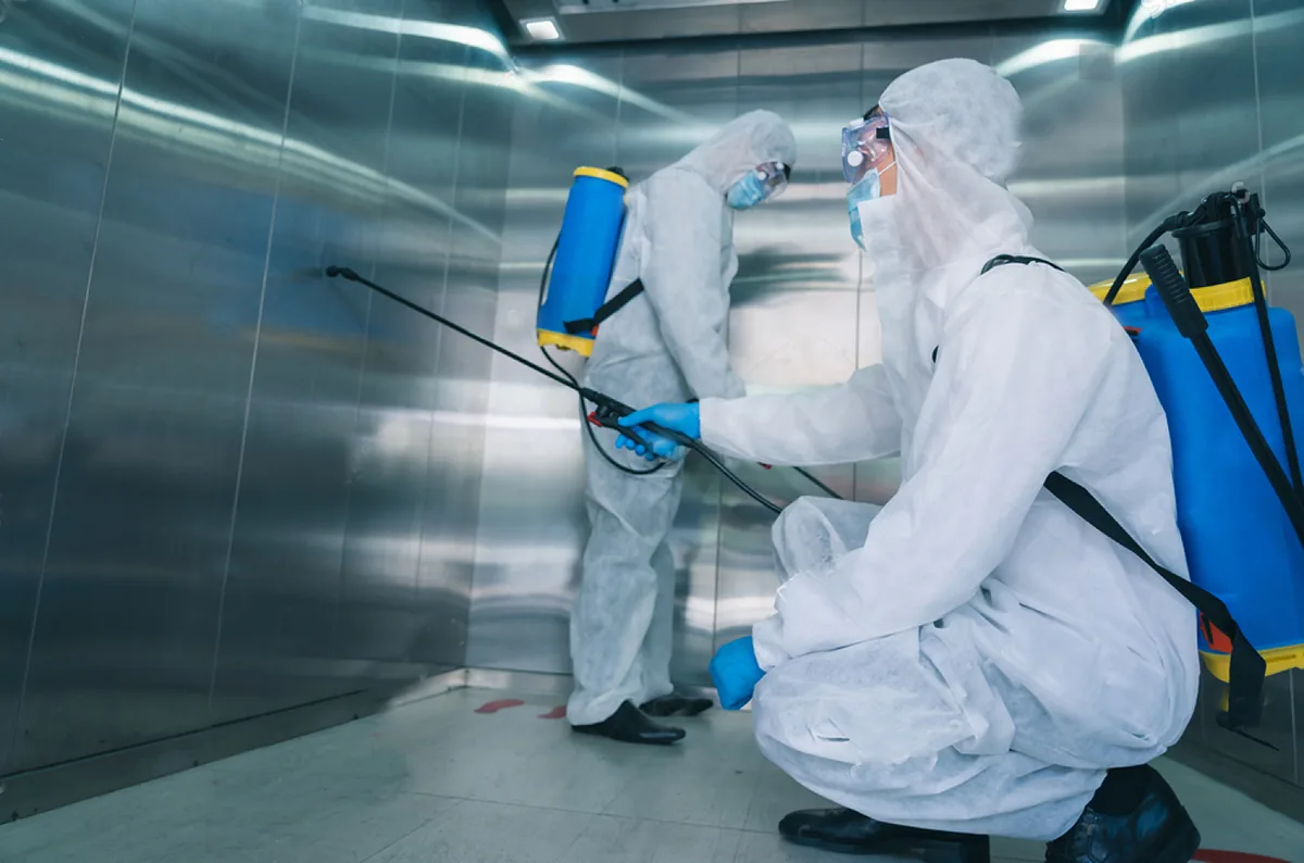 Two workers in white protective suits with sprayers disinfecting inside a metal elevator