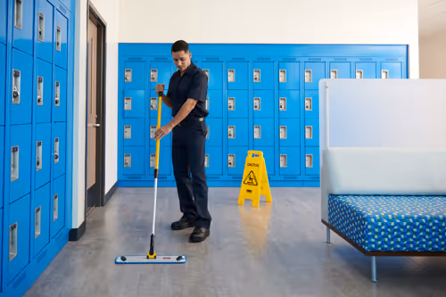 Custodian mopping the floor in a locker room with blue lockers and a caution wet floor sign.