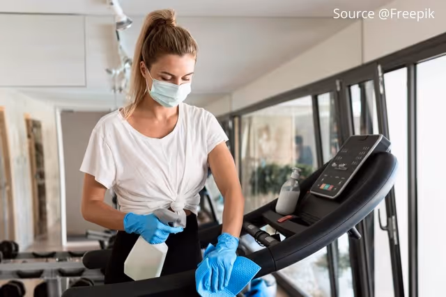 Woman wearing a face mask and gloves cleaning a treadmill with a spray bottle and cloth in a gym.