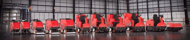 Lineup of red industrial floor cleaning machines inside a large warehouse with metal walls.