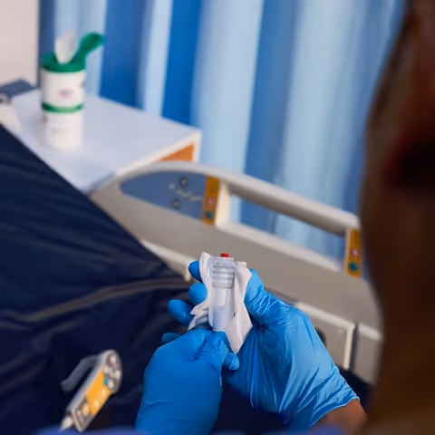 Person wearing blue gloves holding a medical device near a hospital bed with blue curtains in the background.