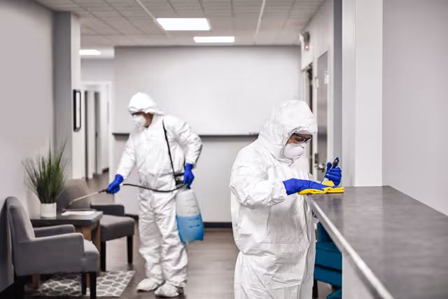 Two workers wearing white protective suits, gloves, masks, and goggles disinfect an indoor space; one sprays a disinfectant while the other wipes a counter with a yellow cloth.