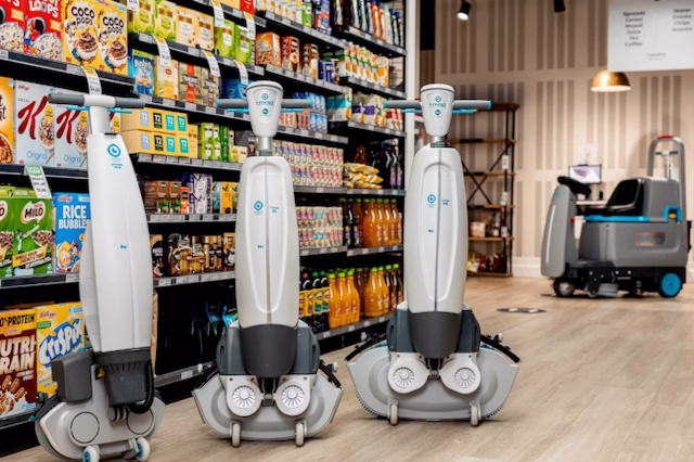 Three white autonomous cleaning robots in front of store shelves stocked with cereals and beverages on a wooden floor.