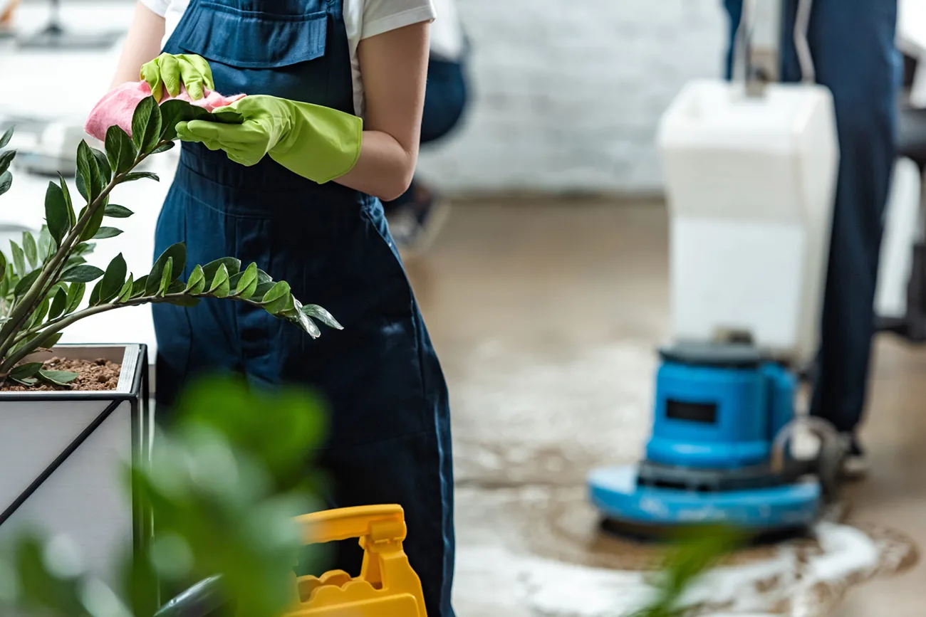 Cleaner in blue overalls and green gloves beside a plant and a floor polishing machine