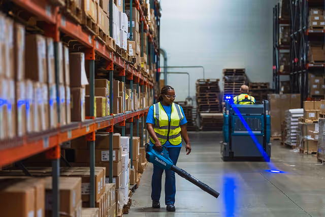 Warehouse worker in a yellow safety vest using a leaf blower to clean the floor between shelves filled with boxes, while another worker drives a floor cleaning machine in the background.