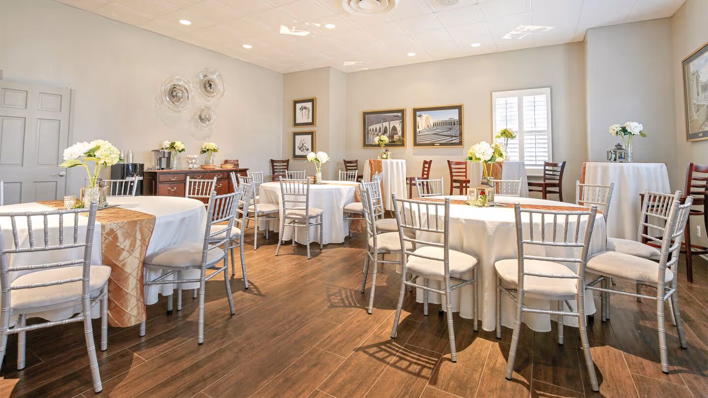 Bright, elegant event room with round tables covered in white and peach tablecloths, silver chairs, and floral centerpieces.