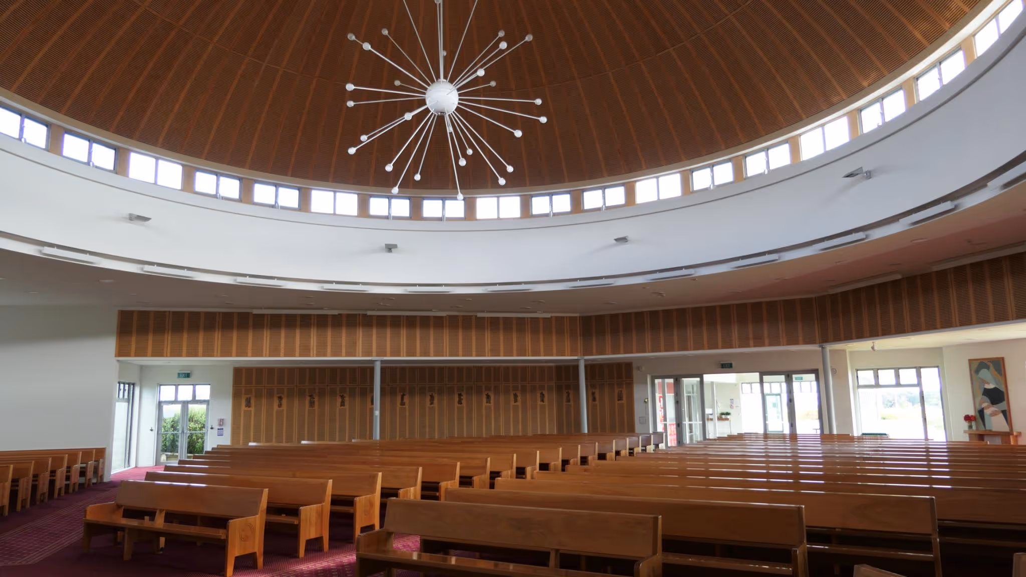 Interior view of a spacious church with wooden pews, a circular wooden ceiling with small windows around the top, and a modern starburst chandelier.